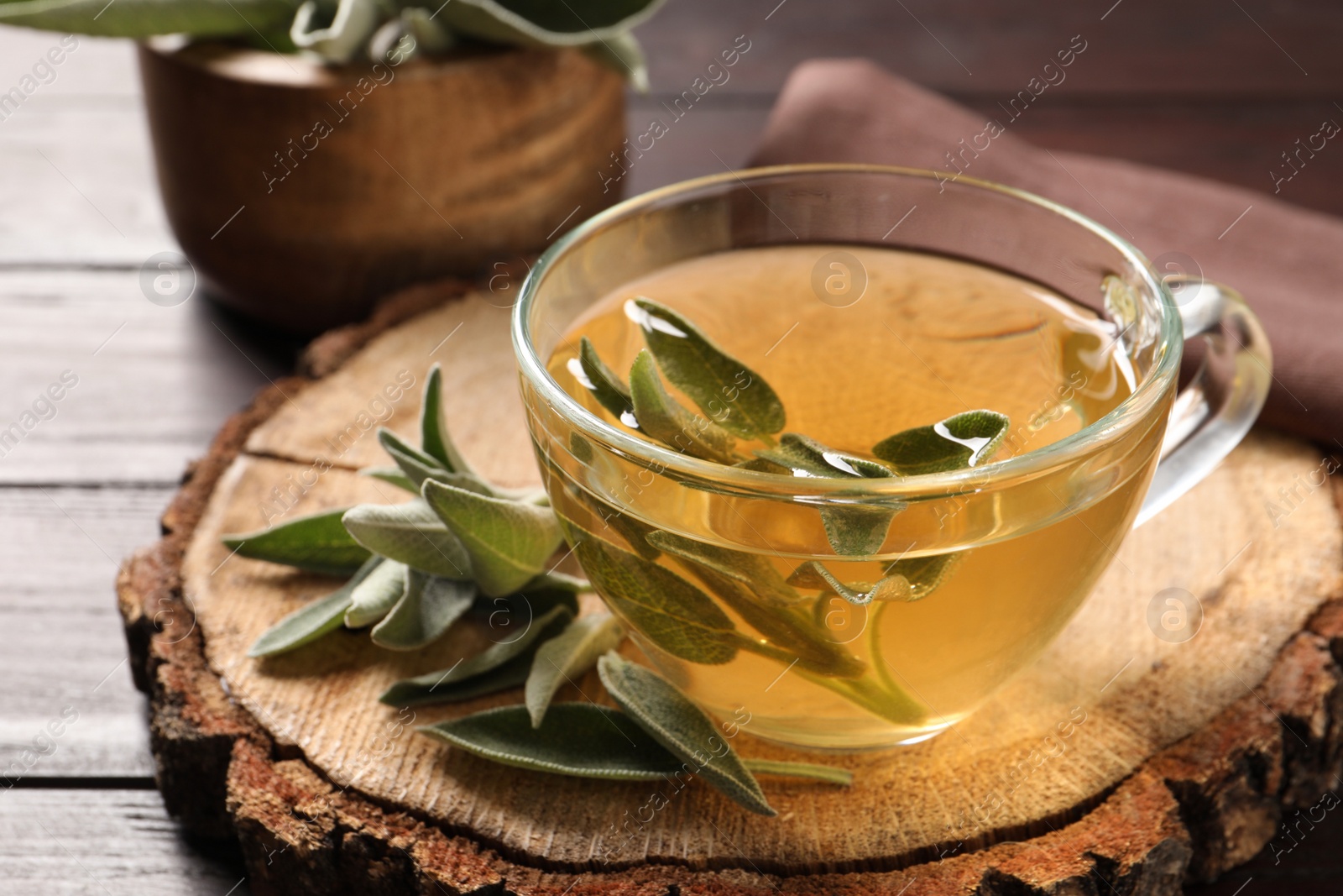 Photo of Cup of aromatic sage tea and fresh leaves on wooden stump, closeup