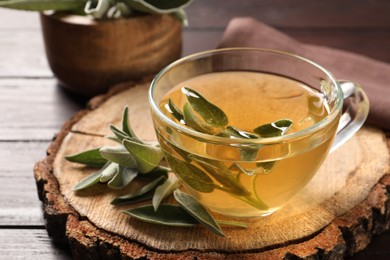 Photo of Cup of aromatic sage tea and fresh leaves on wooden stump, closeup