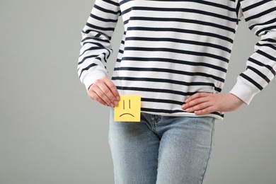 Photo of Cystitis. Woman holding sticky note with drawn sad face on grey background, closeup. Space for text