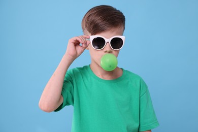 Photo of Boy in sunglasses blowing bubble gum on light blue background