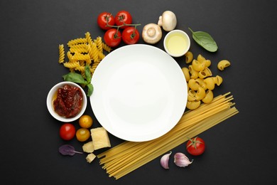 Plate surrounded by different types of pasta and products on black background, flat lay. Space for text