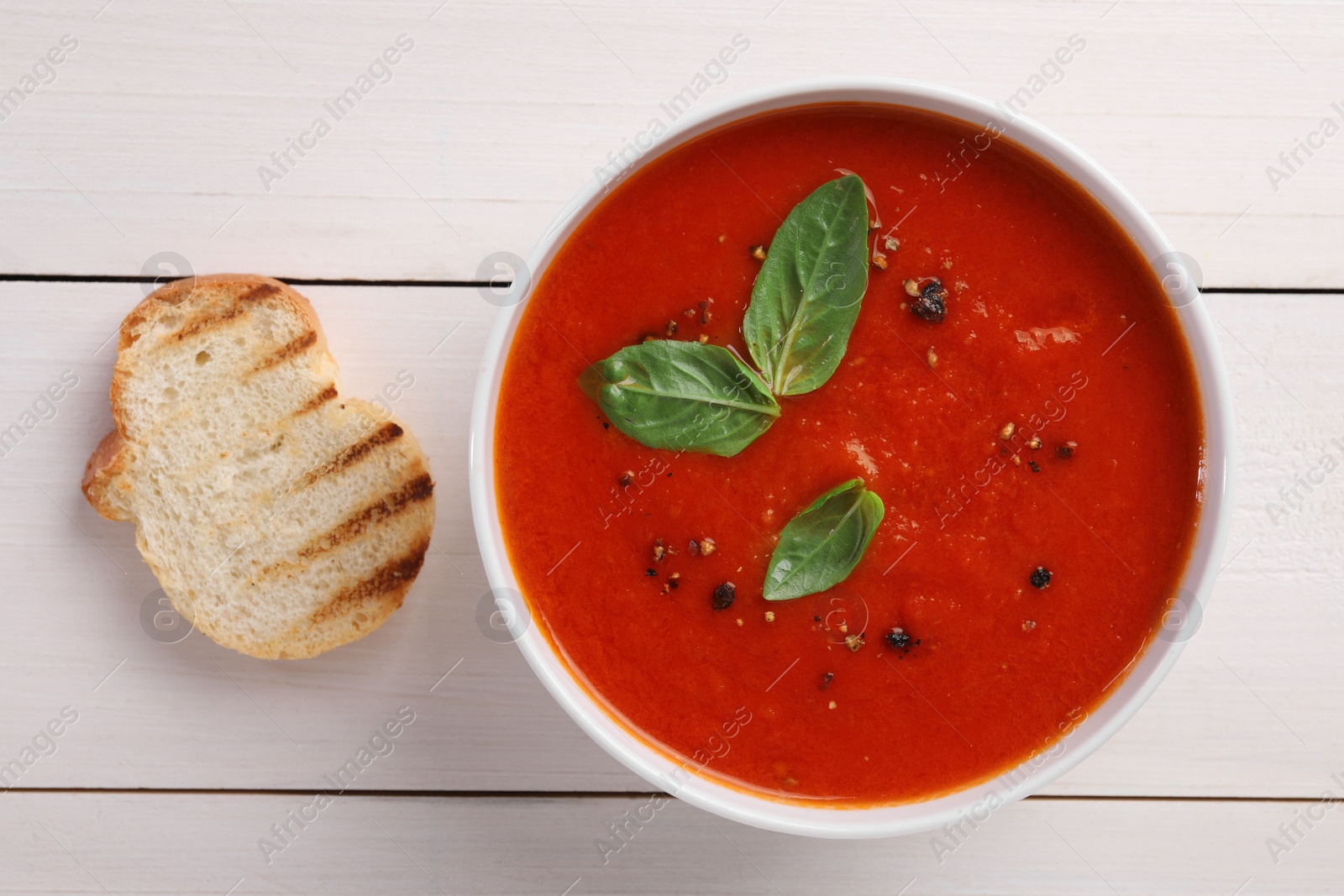 Photo of Delicious tomato cream soup in bowl and piece of grilled bread on white wooden table, flat lay