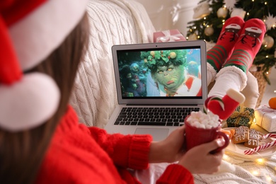 Photo of MYKOLAIV, UKRAINE - DECEMBER 25, 2020: Woman with sweet drink watching The Grinch movie on laptop at home, closeup. Cozy winter holidays atmosphere