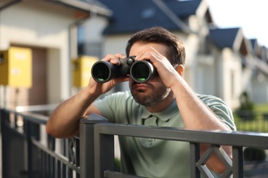 Concept of private life. Curious man with binoculars spying on neighbours over fence outdoors