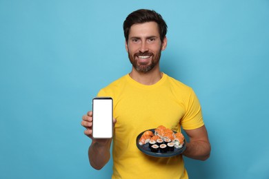 Photo of Happy man holding plate with tasty sushi rolls and smartphone on light blue background