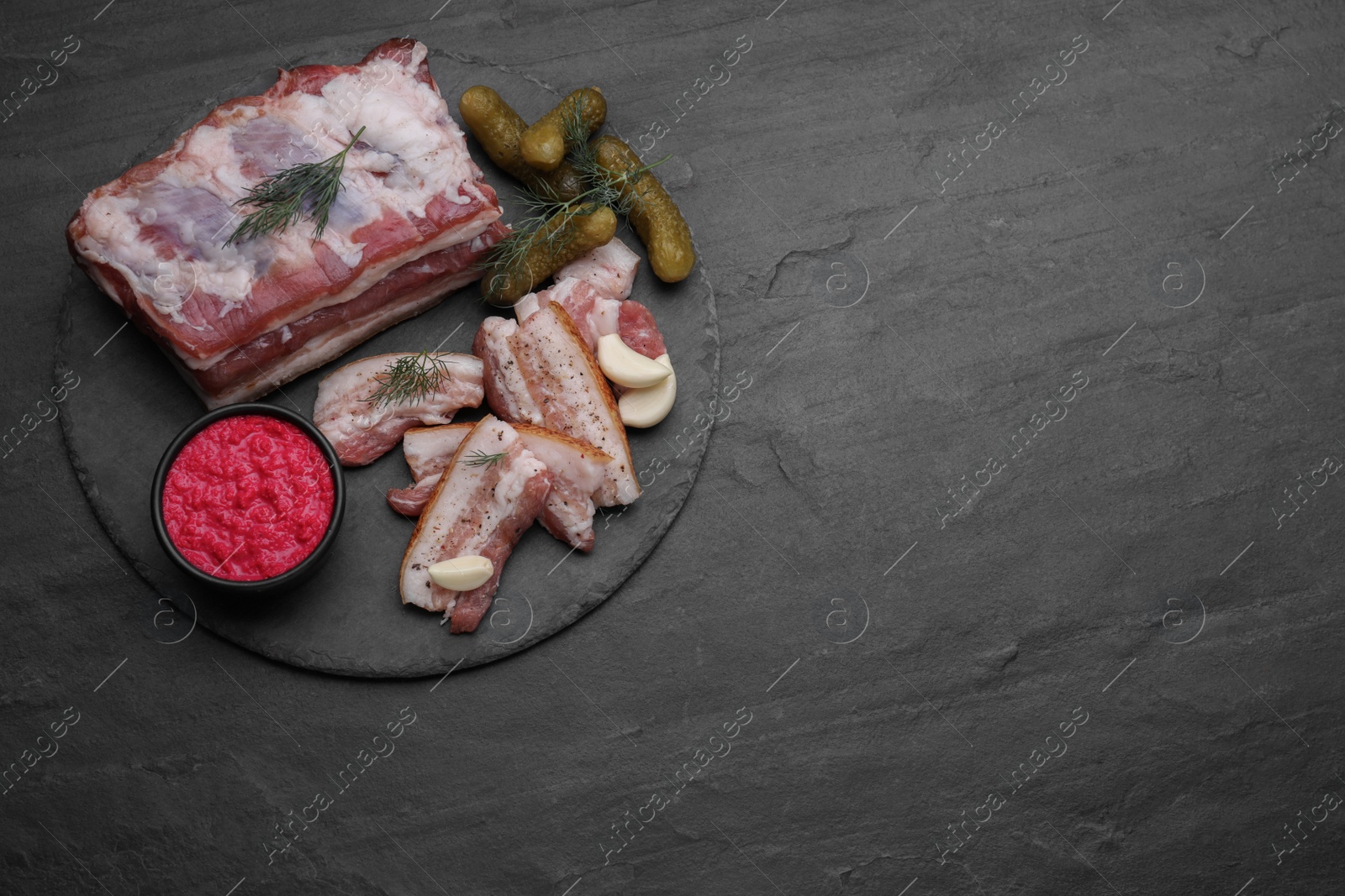 Photo of Tasty pork fatback with spices, pickled cucumbers and sauce on black table, top view. Space for text