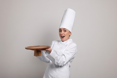 Happy professional confectioner in uniform holding empty cake stand on light grey background