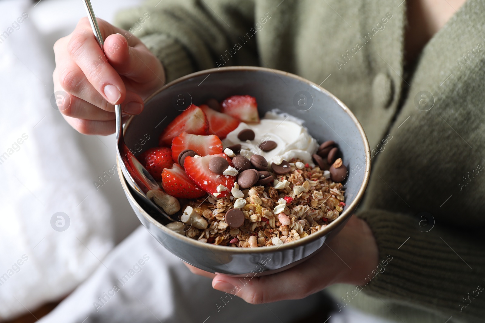 Photo of Woman eating tasty granola with chocolate chips, strawberries and yogurt indoors, closeup