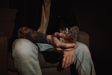 Photo of Young man with tattoos and glass of whiskey sitting in armchair on dark background, closeup