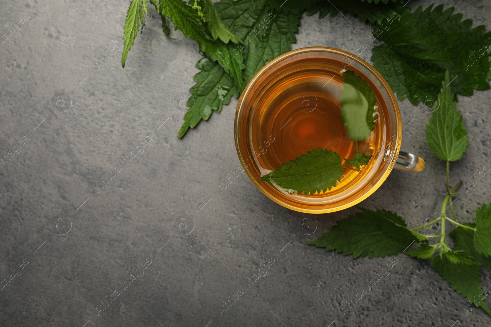 Photo of Glass cup of aromatic nettle tea and green leaves on grey table, flat lay. Space for text