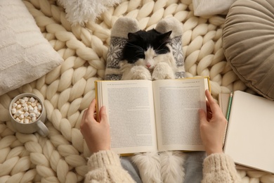 Photo of Woman reading book and holding adorable cat on knitted blanket, top view