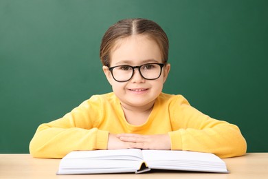 Happy little school child sitting at desk with book near chalkboard