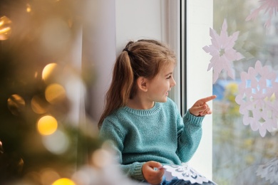 Cute little girl with paper snowflake near window indoors