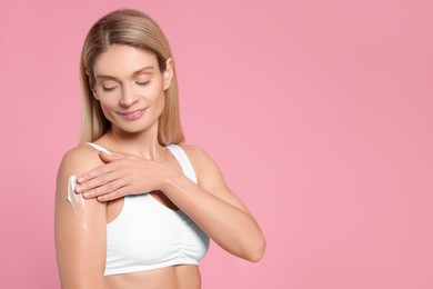 Woman applying body cream onto her arm against pink background, space for text