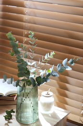 Vase with fresh eucalyptus branches on table near window in room. Interior design
