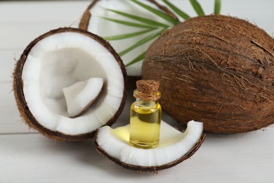 Bottle of organic coconut cooking oil, leaf and fresh fruits on white wooden table, closeup