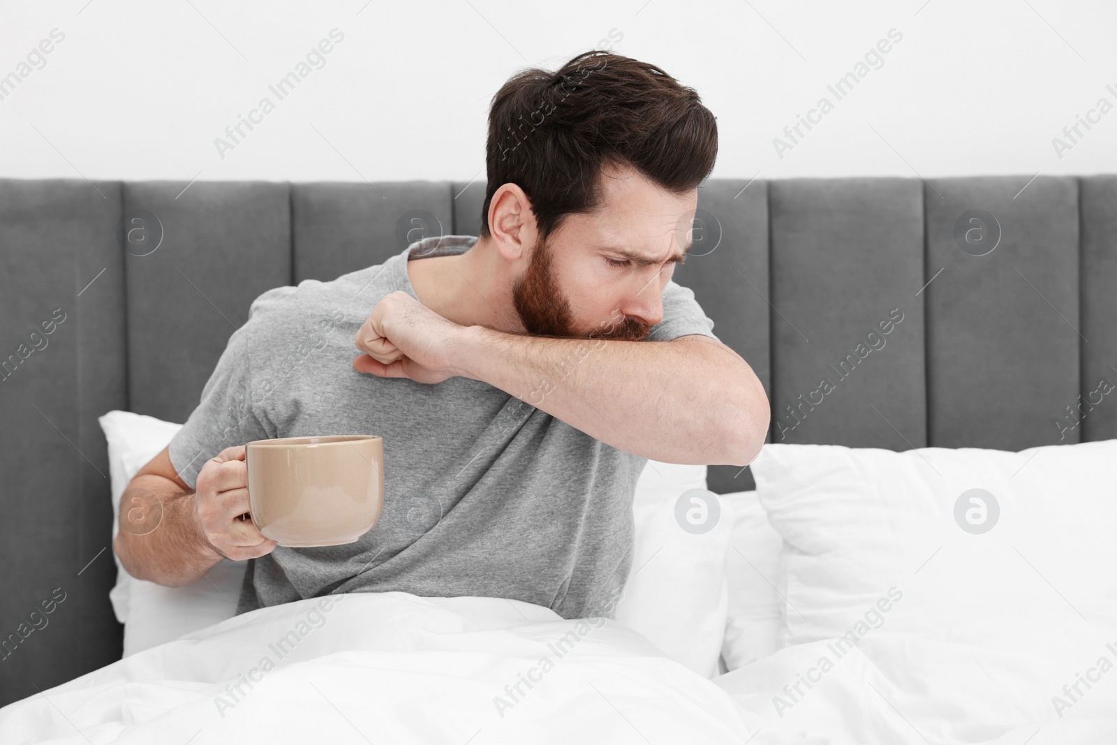 Photo of Sick man with cup of hot drink coughing on bed at home. Cold symptoms