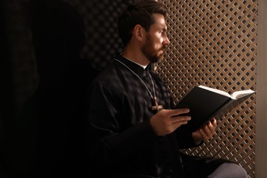 Photo of Catholic priest in cassock reading Bible in confessional booth