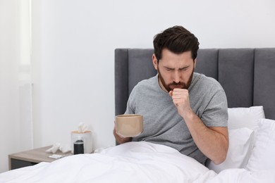 Photo of Sick man with cup of hot drink coughing on bed at home. Cold symptoms