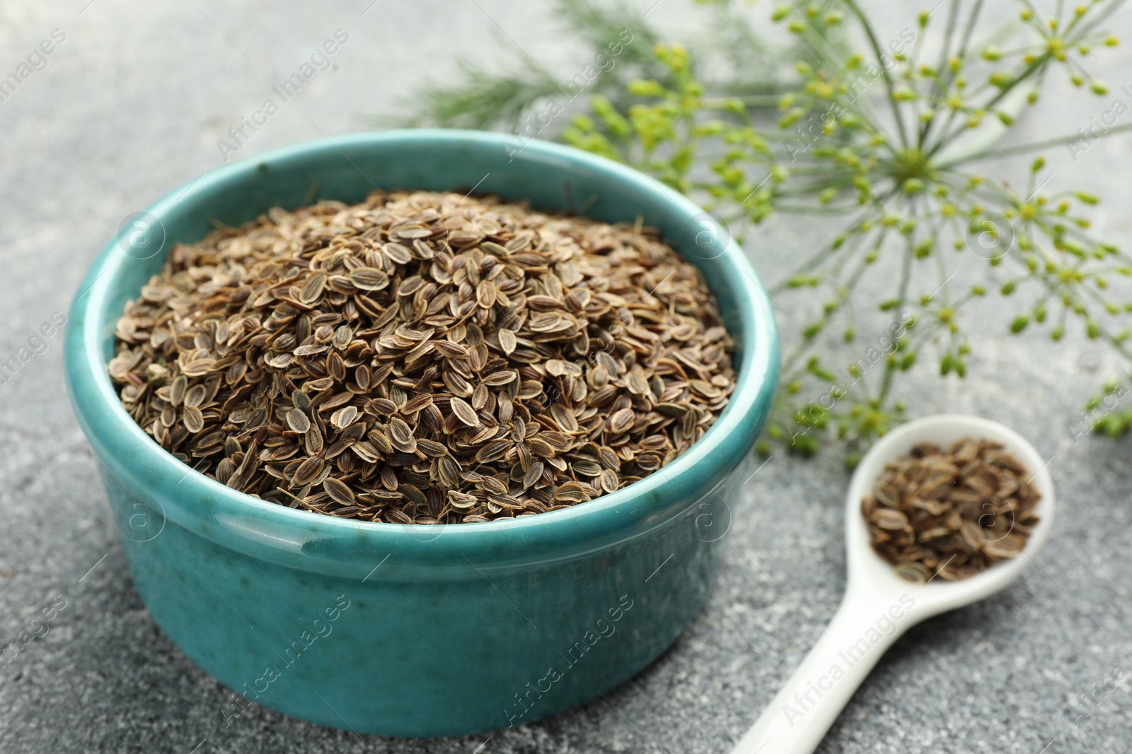 Photo of Bowl of dry seeds, spoon and fresh dill on grey table, closeup