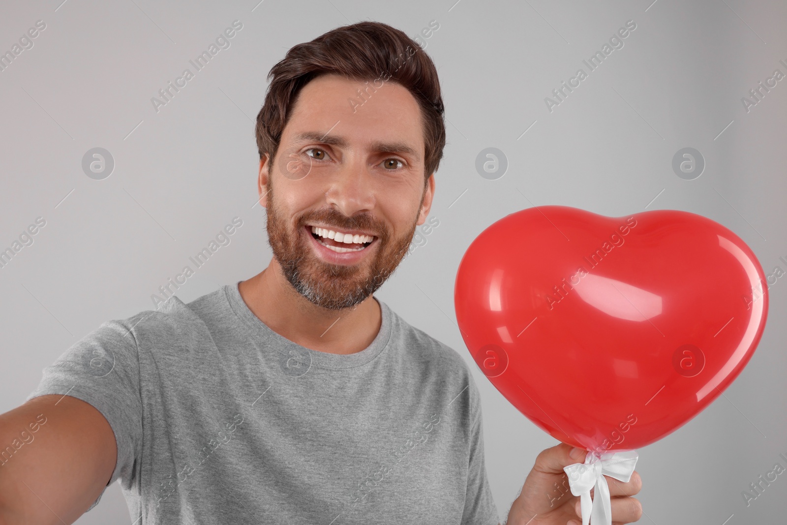 Photo of Happy man holding red heart shaped balloon and taking selfie on grey background