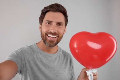 Happy man holding red heart shaped balloon and taking selfie on grey background