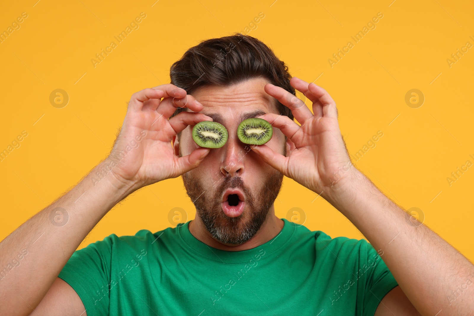 Photo of Emotional man holding halves of kiwi near his eyes on orange background