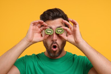 Emotional man holding halves of kiwi near his eyes on orange background