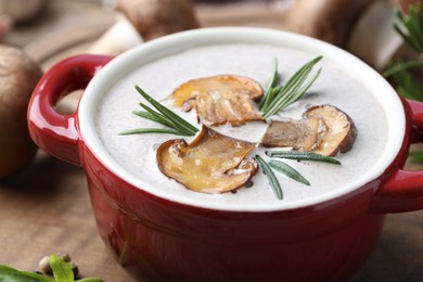 Photo of Delicious homemade mushroom soup in ceramic pot and fresh ingredients on wooden table, closeup