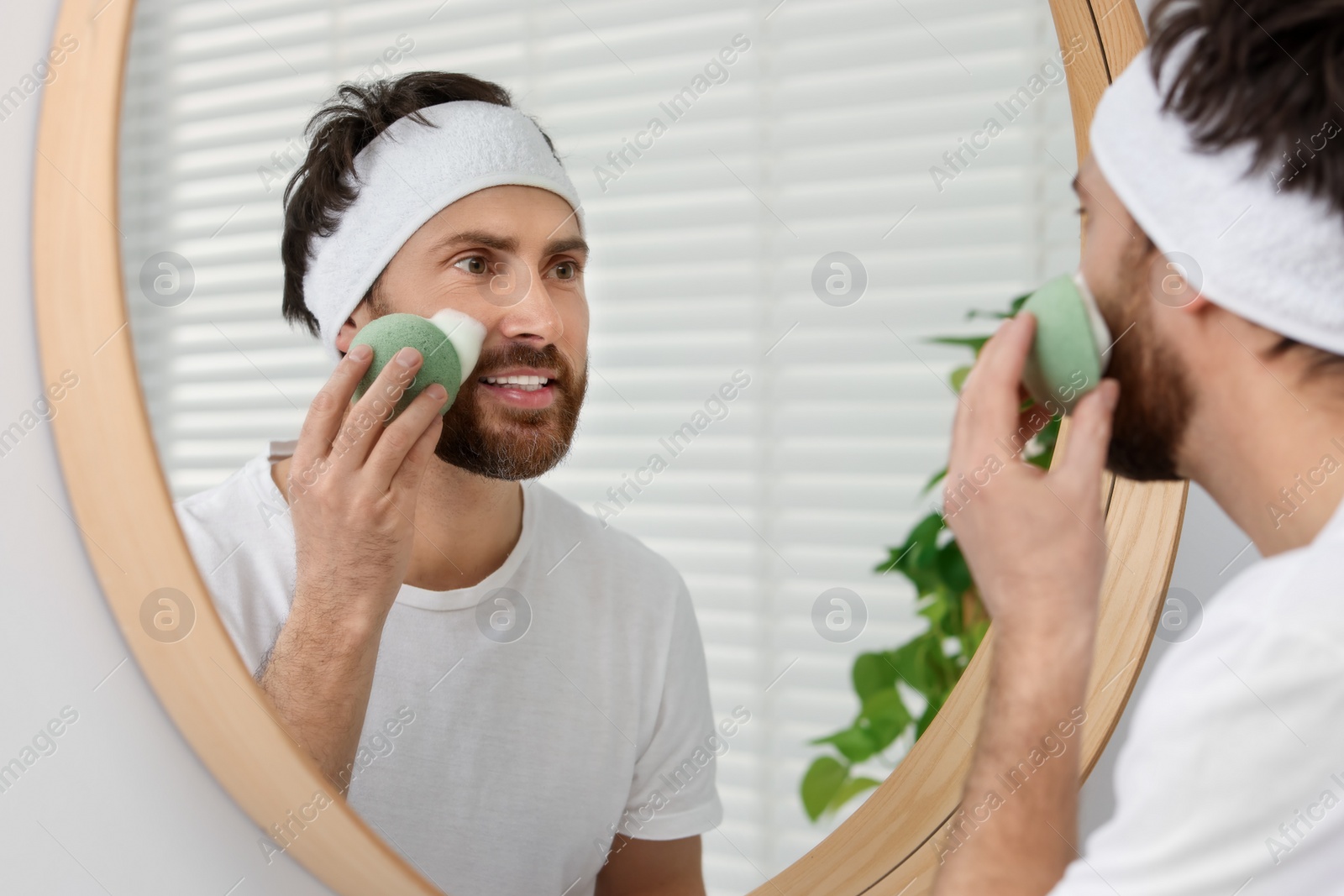 Photo of Man with headband washing his face using sponge near mirror in bathroom