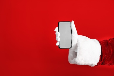 Photo of Santa holding modern mobile phone with blank screen on red background, closeup. Space for text