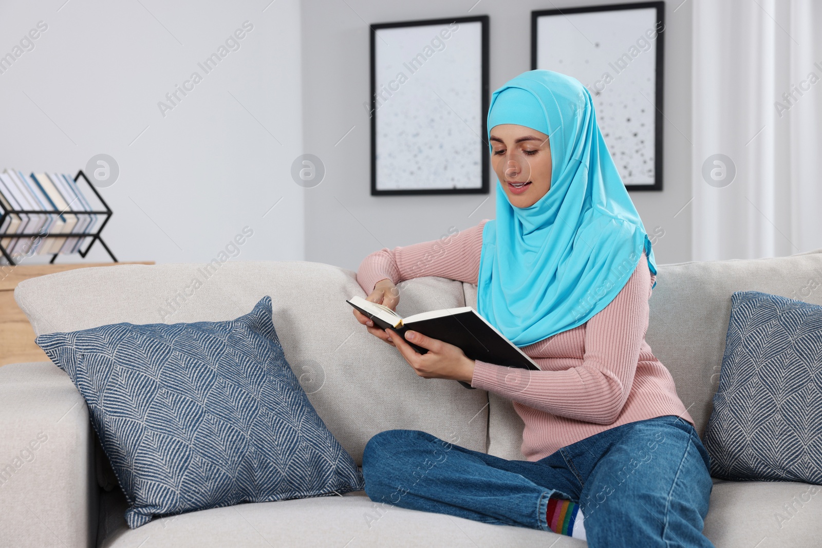 Photo of Muslim woman reading book on couch in room. Space for text