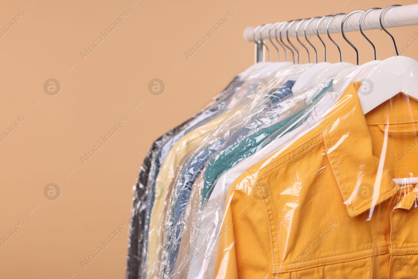 Photo of Dry-cleaning service. Many different clothes in plastic bags hanging on rack against beige background, space for text