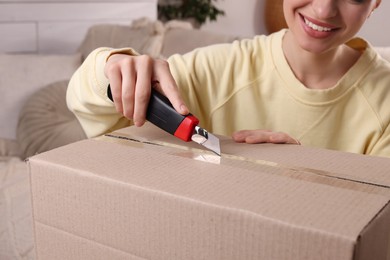 Young woman using utility knife to open parcel at home, closeup