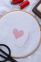 Photo of Embroidered red heart, needle and scissors on white cloth, top view