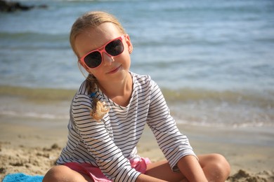 Little girl in stylish sunglasses on sandy beach near sea