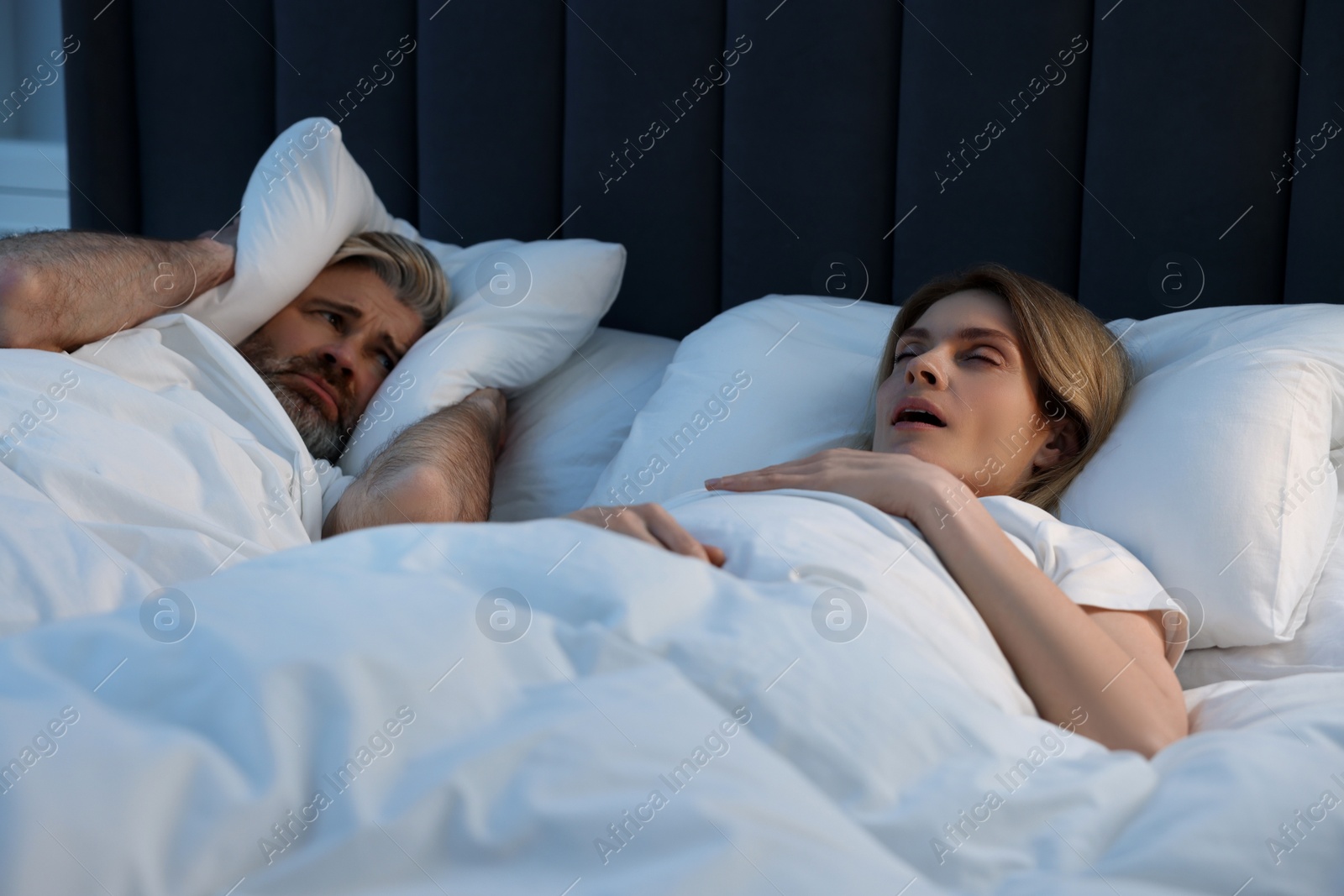 Photo of Irritated man covering his ears with pillows in bed at home. Problem with snoring wife