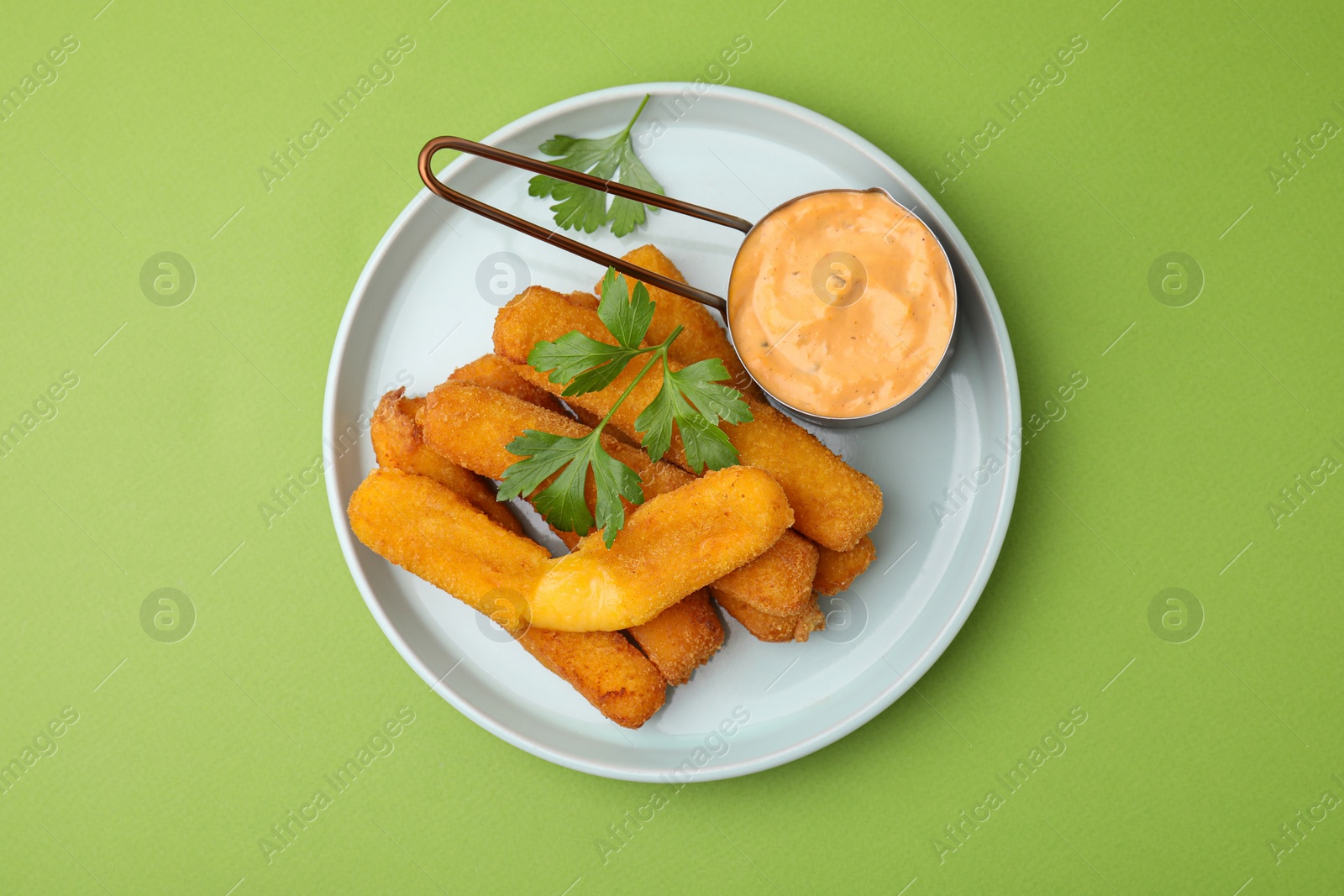 Photo of Tasty fried mozzarella sticks served with sauce and parsley on green background, top view