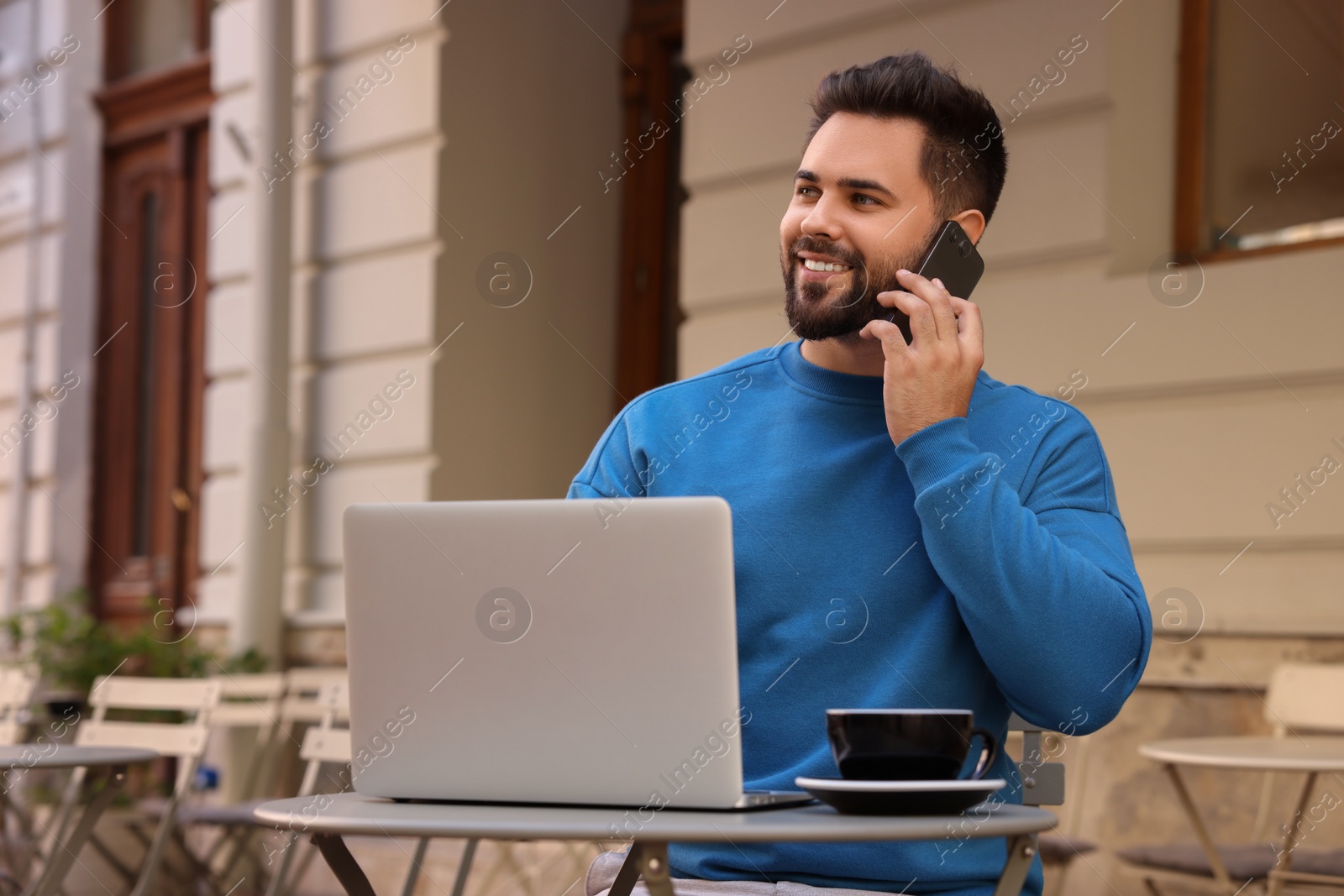 Photo of Handsome young man talking on smartphone while using laptop at table in outdoor cafe