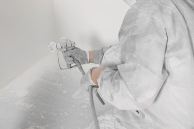 Decorator in protective overalls painting wall with spray gun indoors, closeup