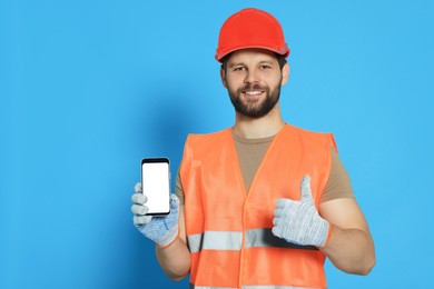 Man in reflective uniform showing smartphone and thumbs up on light blue background, space for text