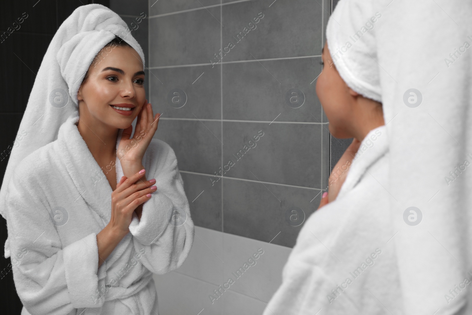 Photo of Beautiful young woman in bathrobe with towel on head near mirror at home