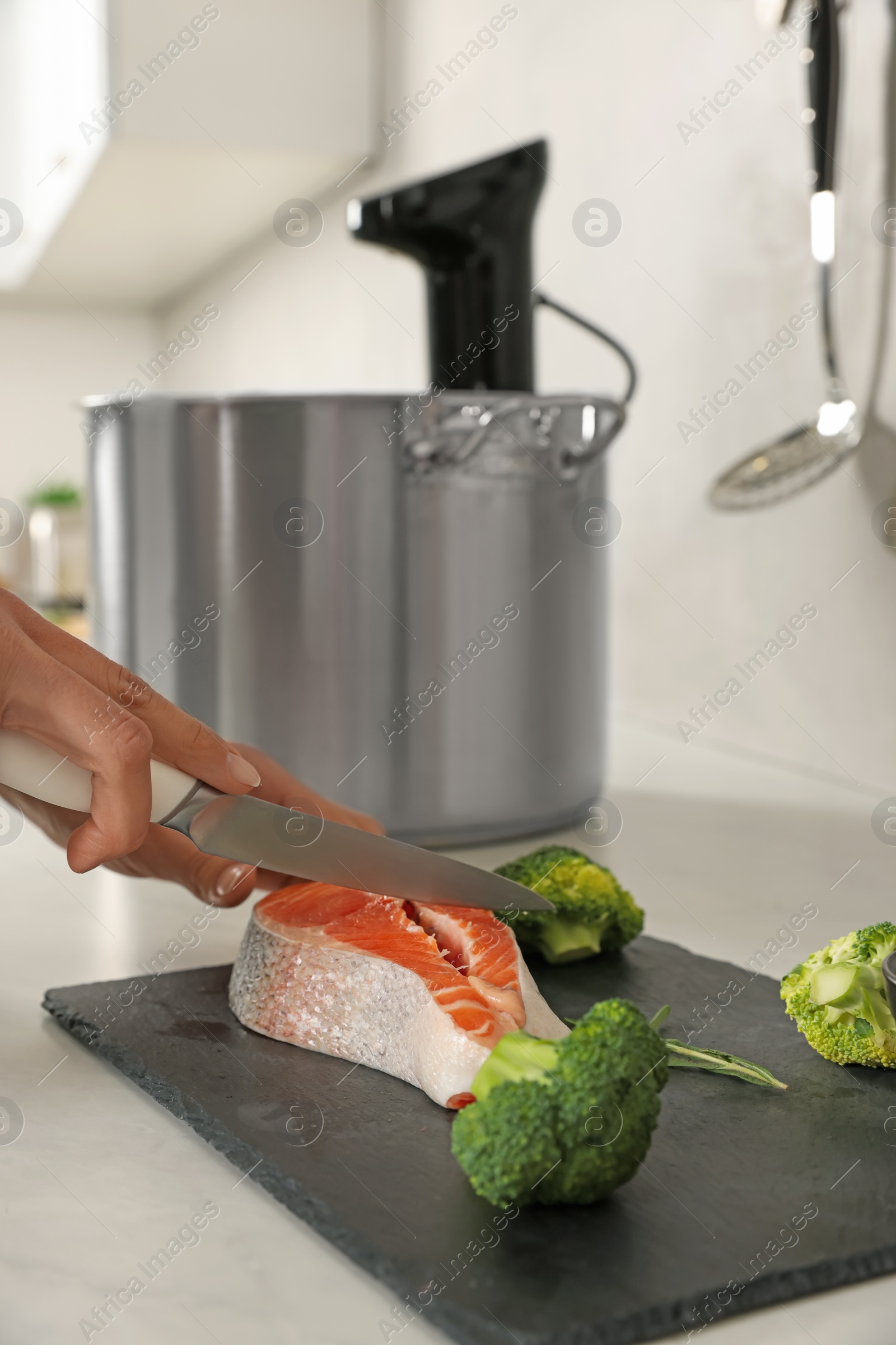 Photo of Woman cutting salmon near pot with sous vide cooker in kitchen. Thermal immersion circulator