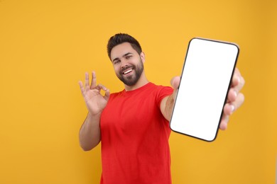 Photo of Young man showing smartphone in hand and OK gesture on yellow background