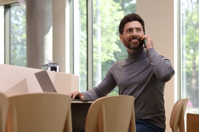 Photo of Handsome man talking on smartphone while using laptop at table in cafe