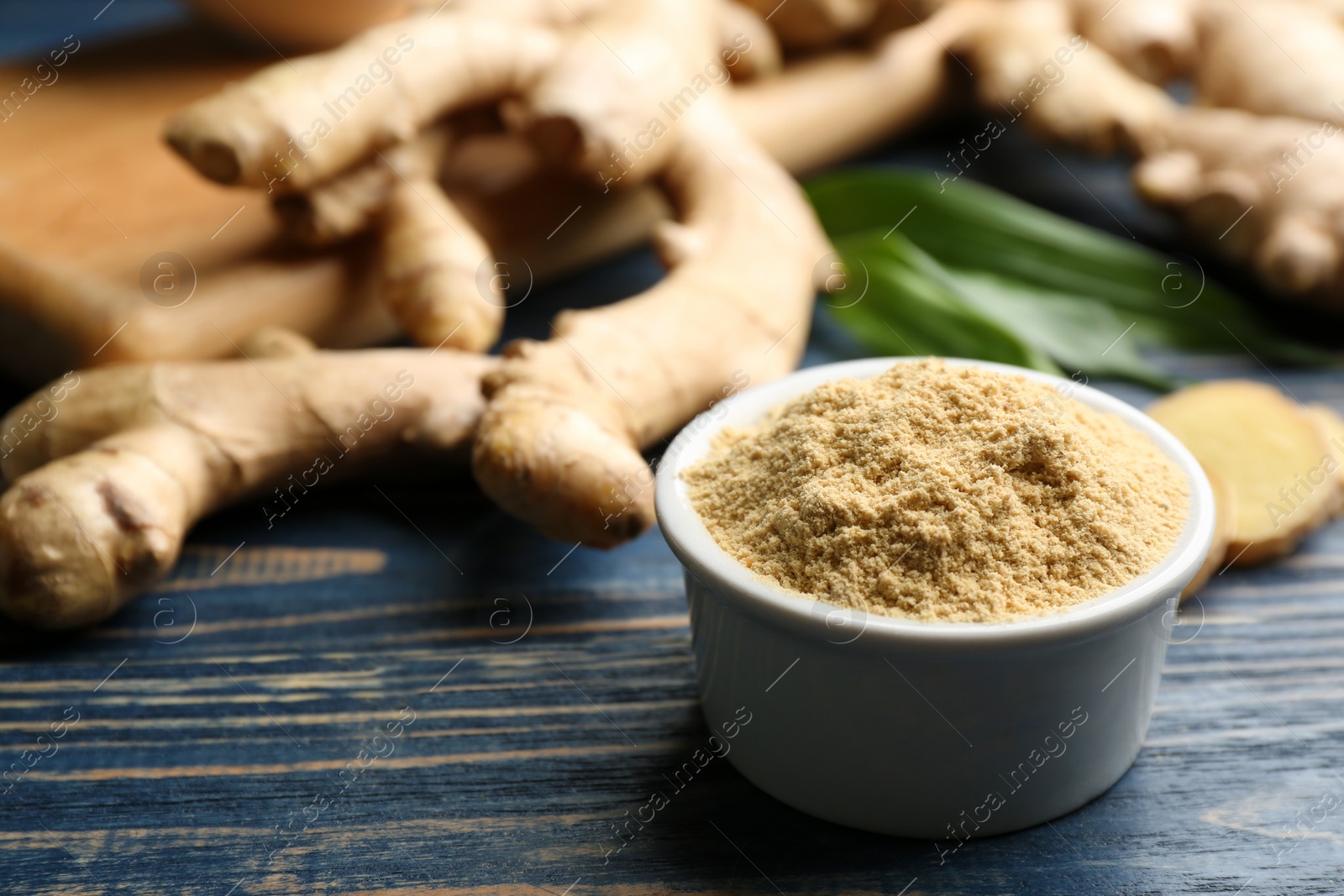 Photo of Dry ginger powder in bowl and fresh root on blue wooden table