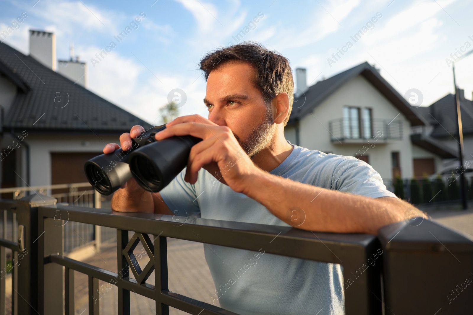 Photo of Concept of private life. Curious man with binoculars spying on neighbours over fence outdoors