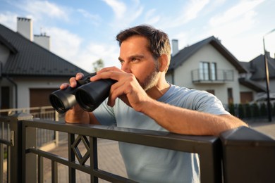 Photo of Conceptprivate life. Curious man with binoculars spying on neighbours over fence outdoors