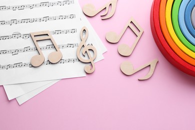 Photo of Baby songs. Music sheets, wooden notes and toy rainbow on pink background, flat lay