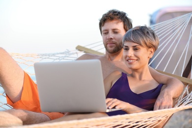 Young couple resting with laptop in hammock on beach